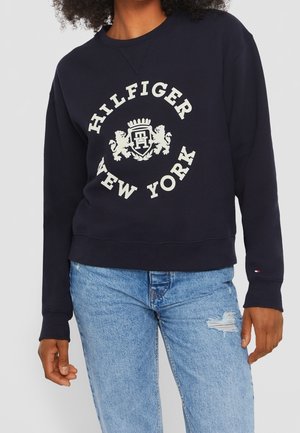 Osoba nosząca granatową bluzę z napisem i emblematem „Hilfiger New York”, w zestawieniu z jasnoniebieskimi, podartymi dżinsami.