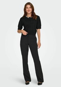 Juoda trumpa megztinė su trumpomis rankovėmis, apvali apykaklė, papuošta dekoratyviniais akcentais, derinama su tiesiomis juodomis kelnėmis ir slip-on batais.