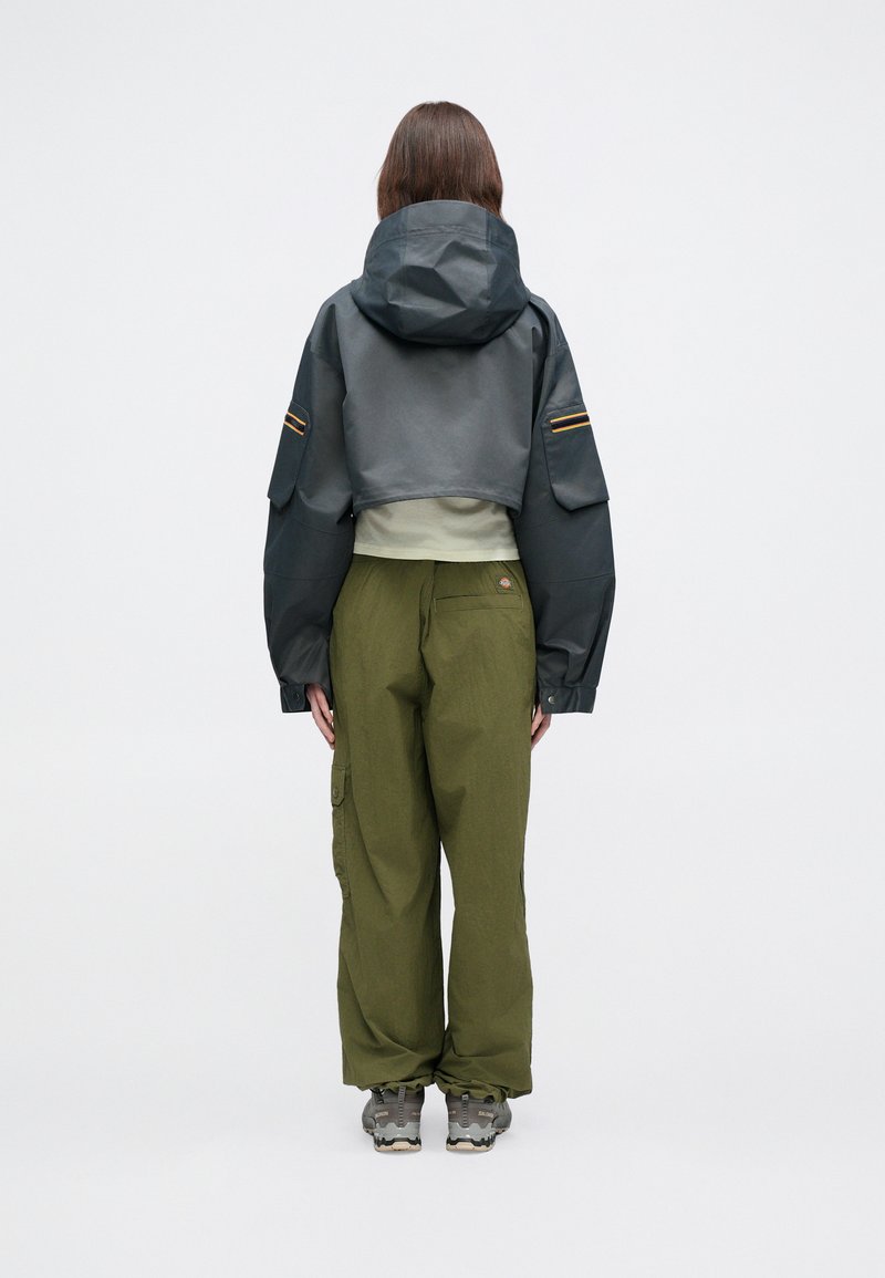 Krajša siva vodoodbojna jakna s kapuco, kombinirana z ohlapnimi olivno zelenimi cargos hlačami in sivimi pohodnimi čevlji. Ponaša se z žepi in kontrastnimi črtami.