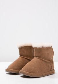 Brązowe zamszowe botki za kostkę z wewnętrzną podszewką z owczej wełny, okrągłym noskiem oraz płaską gumową podeszwą, z detalami szwów na zewnętrznej stronie.