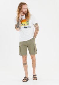 Witte T-shirt met een kleurraf beach graphic, olijfgroene cargoshorts met zijzakken, bruine sandalen en zichtbare tatoeages op armen en benen.