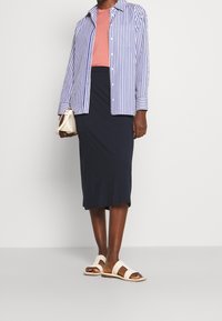 Camicia a righe blu e bianche con bottoni, t-shirt corallo, gonna a matita blu navy e sandali beige. Include una piccola borsa bianca. Tessuti lisci.