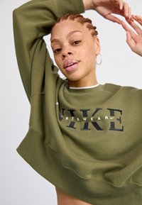 Maslinasto zeleni Nike dukserica s opuštenim fitom, rebrastim manžetama i velikim crnim grafičkim natpisom sprijeda. Mekana tekstura s širokim izrezom.