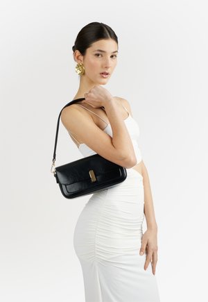 Donna che indossa un vestito bianco arricciato e orecchini dorati tiene una borsa a tracolla nera in pelle con una fibbia dorata sulla spalla.
