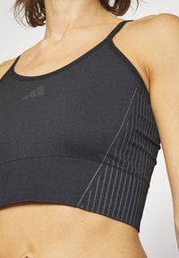 Crna sportska majica izrađena od rastezljivog materijala, s rebrastom teksturom, tankim trakama i dijagonalnim prugastim detaljima sa strane.