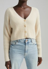Krémově zbarvený krátký kardigan s výstřihem do V, žebrovanou texturou a třemi knoflíky vpředu. Kombinován s světle modrými džínami.