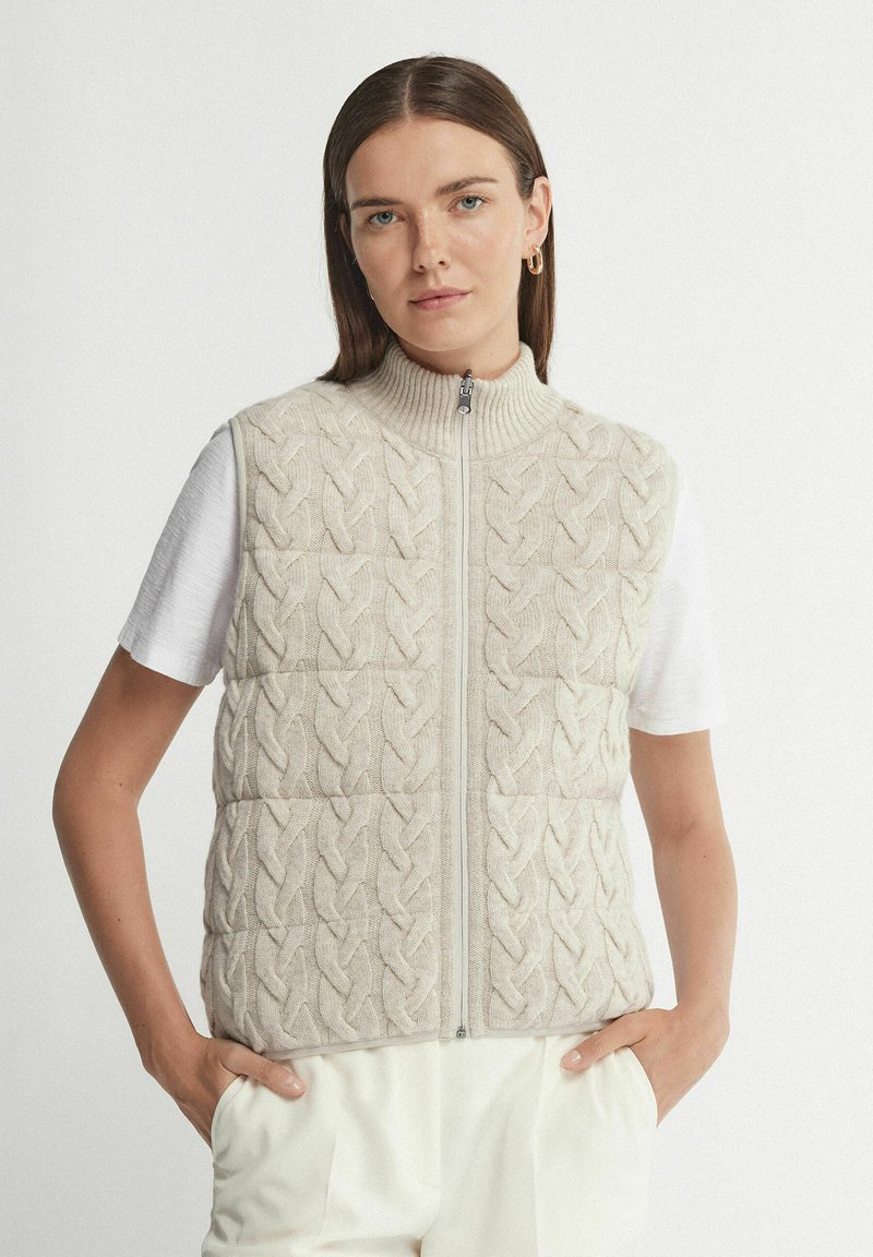Jonge vrouw draagt een beige kabelgebreid vest met ritssluiting over een wit shirt met korte mouwen, haar handen in de zakken van witte broek.