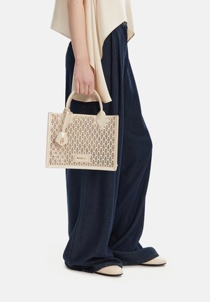 Person trägt weite marineblaue Hosen und beige Schuhe und hält eine beige perforierte Tragetasche mit geometrischen Mustern. 