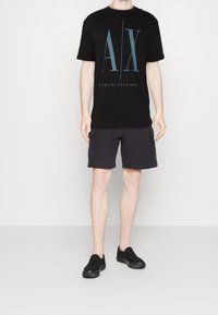 Man som bär en svart Armani Exchange T-shirt, mörka cargo-shorts och svarta slip-on skor, stående mot en ljus enfärgad bakgrund.