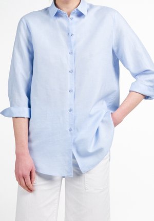 Persona che indossa una camicia azzurro chiaro a maniche lunghe con le maniche arrotolate e pantaloni bianchi, in piedi con una mano in tasca.