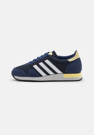 Scarpe sportive blu navy con strisce bianche, dotate di tomaia texturizzata, colletto imbottito e un accento giallo sul tallone. Suola in gomma.