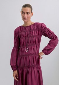 Bordeaux, jurk met lange mouwen en gerimpelde details in een ontspannen silhouet. Voorzien van een ronde halslijn en een vloeiende rok. Accentuering met een zilveren ronde ketting.