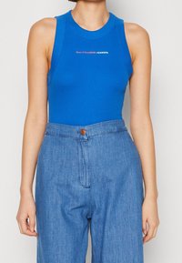 Blå ärmlös bodysuit med rund nacke, prydd med en tryckt logotyp i vitt och rött. Kombinerad med högmidjade denimbyxor.