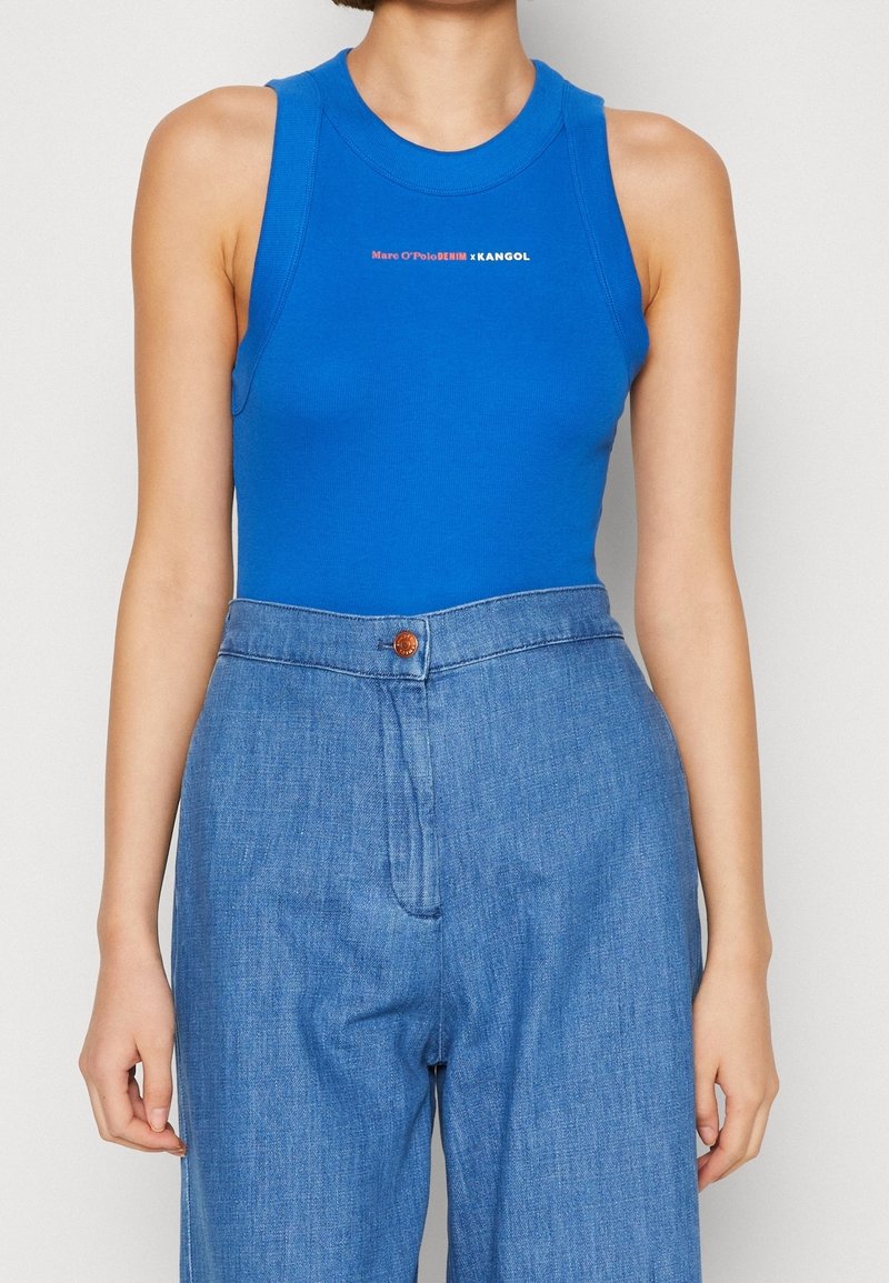 Blå ärmlös bodysuit med rund nacke, prydd med en tryckt logotyp i vitt och rött. Kombinerad med högmidjade denimbyxor.