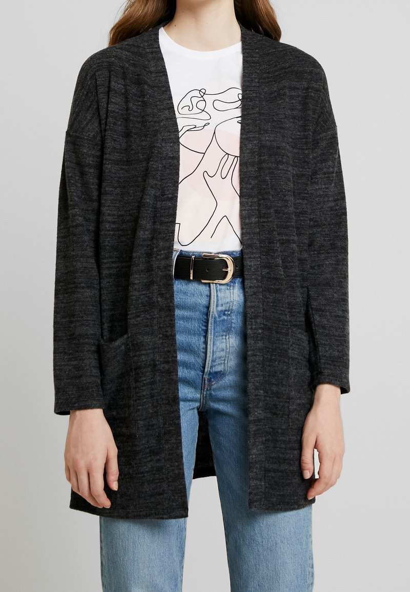 Personne portant un cardigan gris foncé ouvert devant, un t-shirt blanc à motif, un jean bleu et une ceinture noire avec une boucle dorée sur un fond uni.