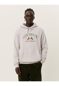 Felpa grigia con cappuccio regolabile, tasca a marsupio frontale e logo stampato "LES DEUX". Indossata con pantaloni in velluto a coste verde scuro.