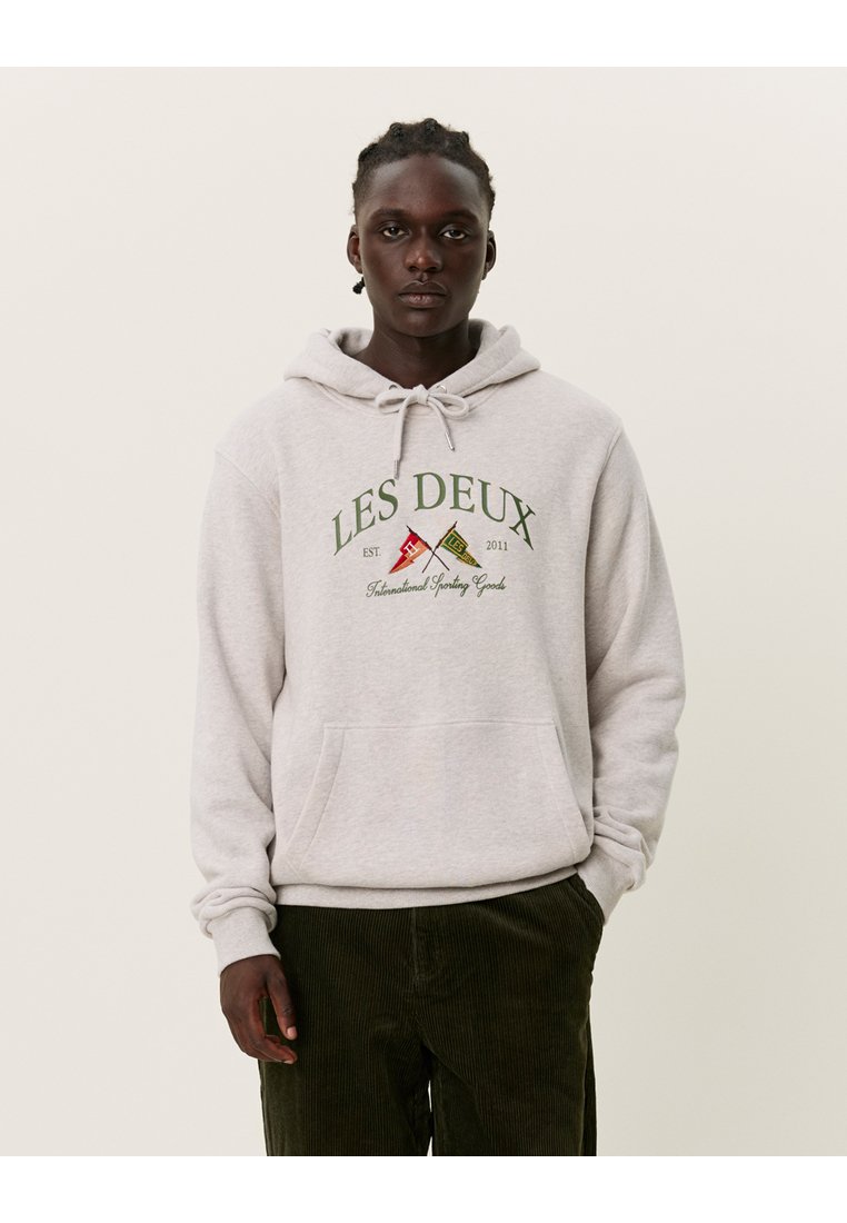 Sweatshirt cinza com capuz ajustável, bolso frontal tipo pouch e logótipo impresso "LES DEUX". Usado com calças de corduroy verde-escuras.