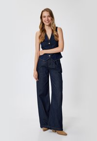 Denim-puku, joka sisältää tummansinisen, henkselittömän liivin, jossa on napit ja leveälahkeiset housut. Pehmeä rakenne ja muotoiltu istuvuus.