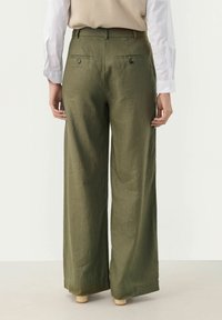 Pantaloni a gamba larga in lino verde oliva, con tasche posteriori con bottoni e una texture liscia, indossati con un top beige e maniche di camicia bianche.