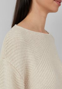 Donna con lunghi capelli scuri che indossa un maglione lavorato a maglia color crema con un collo ampio e arrotondato, su uno sfondo grigio semplice.