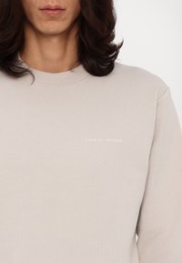 Jasnobeżowa bluza crewneck wykonana z miękkiego materiału, z delikatnym logo "TIGER OF SWEDEN" w jasnym nadruku na piersi.