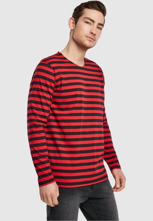 Mann, der ein rot-schwarz horizontal gestreiftes Langarmshirt und dunkle Jeans trägt, steht vor hellgrauem Hintergrund.