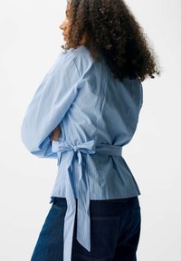 Camicia avvolgente a strisce blu chiaro con maniche lunghe, caratterizzata da una vita legata e una texture liscia. Abbinata a jeans in denim scuro.