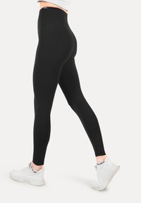 Zwarte high-waisted leggings van rekbaar stof, met een gladde afwerking en een getailleerd ontwerp, gecombineerd met witte sportschoenen.