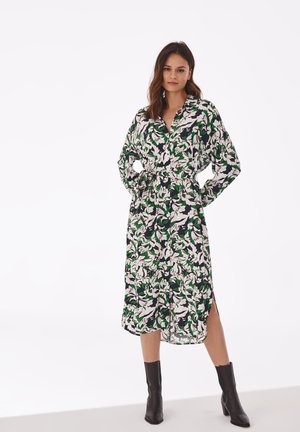 Langes Hemdkleid mit schwarzem Hintergrund, das mit weißen und grünen Blumenmustern verziert ist, mit Knopfleiste und seitlichen Schlitzen. Getragen mit schwarzen Knöchelstiefeln.