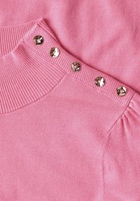 Gros plan de l'épaule d'un pull rose en tricot montrant un col côtelé et cinq boutons ronds brillants le long de la couture.