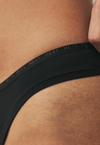Mutande nere con una consistenza liscia e una vita ampia e piatta che presenta un sottile branding "NEXT". Inquadratura ravvicinata del lato e della pelle.