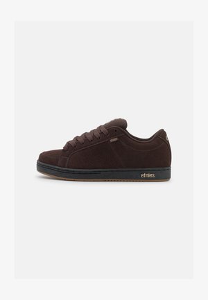 Mørkebrune mokasiner i semsket skinn med avrundet tå, perforerte sidelommer og matchende lisser. Har gummisåle og gull "etnies"-logo på hælen.