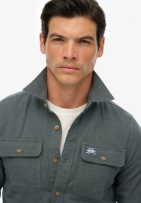 Man met donker haar die een grijs overhemd met klepzakken en een witte onderhemd draagt, kijkt recht in de camera.