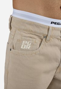 Pantalonii din denim bej prezintă o textură subtilă în zigzag, un buzunar frontal cu logo brodat și un talie albă cu dungi gri.