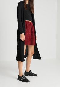 Black ribbed long cardigan over a fitted black crop top, paired with a red suede pleated skirt and black buckle-detail shoes.