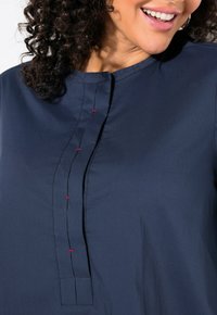 Marineblaue Bluse aus glattem Stoff, mit Stehkragen, Knopfleiste vorne mit dekorativer roter Stichnaht und lockerer Passform.