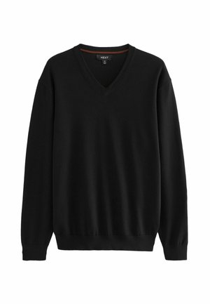 Mann trägt schwarzen V-Ausschnitt Pullover, dunkle Hose und schwarze Sneakers mit weißen Sohlen, posiert mit einer Hand in der Tasche vor grauem Hintergrund.