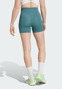 Høyt liv teal shorts i elastisk stoff, med flate sømmer og en jevn finish. Kombinert med hvite sokker og lyse grønne sneakers.