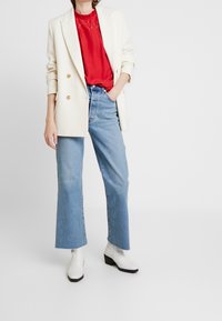 Creme dobbeltradet blazer, rød bluse med blondedetaljer, højtaljede lysblå klokkejeans og hvide ankelstøvler med blokhæle.