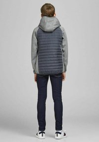 Szara kurtka z kapturem z pikowanym ciemnoszarym tyłem, zestawiona z ciemnymi dżinsami i białymi sneakersami, widziana z tyłu. Gładka tekstura materiału.