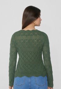Grüner langärmeliger Strickpullover mit einem Rautenmuster und gewelltem Saum, aus strukturiertem Stoff und mit offener Webstruktur, kombiniert mit blauen Jeans.