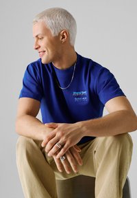 Νεαρός άνδρας με κοντά πλατινένια ξανθά μαλλιά, φοράει μπλε μπλουζάκι Red Bull Racing, μπεζ παντελόνι, ασημένιο κολιέ και δαχτυλίδια, καθισμένος κοιτάζοντας αριστερά.