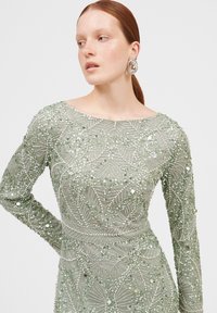 Grünes Paillettenkleid mit langen Ärmeln, das mit aufwendigen Perlen- und Musterdetails verziert ist; runder Halsausschnitt und strukturierter Stoff. Verbundene metallische Ohrringe.