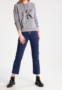 Sudadera gris con un gran logo negro de 'CK' y texto blanco. Combinada con jeans recortados de color azul oscuro y botines negros de cuero charolado.