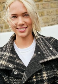 Manteau en laine à carreaux noir et beige avec un grand col et des boutons-pression en argent, porté sur un t-shirt blanc avec un tissu texturé.