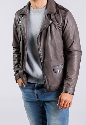 Mann trägt eine dunkelbraune Lederjacke über einem hellgrauen Pullover und blauen Jeans, mit den Händen in den Taschen vor hellem, einfachem Hintergrund.