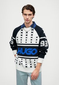 Pleten pulover z črno-belim geometrijskim vzorcem, z modrim logotipom "HUGO", rebrastimi manšeti in slojevito modro srajco s kragnico.