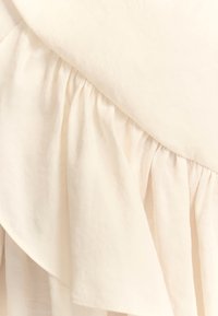 Tissu crème avec une texture douce. Présente un détail de volants superposés le long du bord, créant un design fluide.