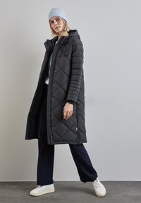 Černý, dlouhý prošívaný kabát s prošívaným designem, se zipovým předním zapínáním a bočními kapsami. Nesený přes tmavě modré kalhoty a bílé tenisky, s světle modrým kulichem.