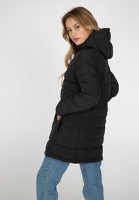 Sort, puffy lang frakke med hætte, der har quiltet design og sidelommer. Sammen med lyseblå brede jeans.